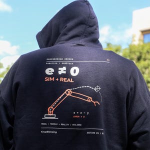 Puede incluir: Una sudadera con capucha azul marino oscuro que presenta un gráfico blanco y naranja en la parte posterior. El diseño incluye el texto "ENGINEERING SERIES", "e ≠ 0", "SIM → REAL" y "MODEL / REALITY", junto a una ilustración técnica de un brazo robótico.