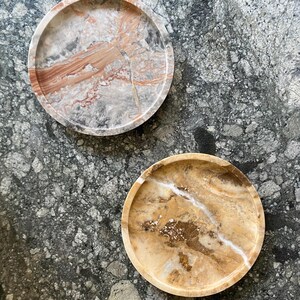 May include: Two circular stone trays with raised edges. The top tray features a marbled pattern of grey, white, and reddish-brown tones. The bottom tray displays a warm amber and golden-brown color palette with white veining and crystalline details.