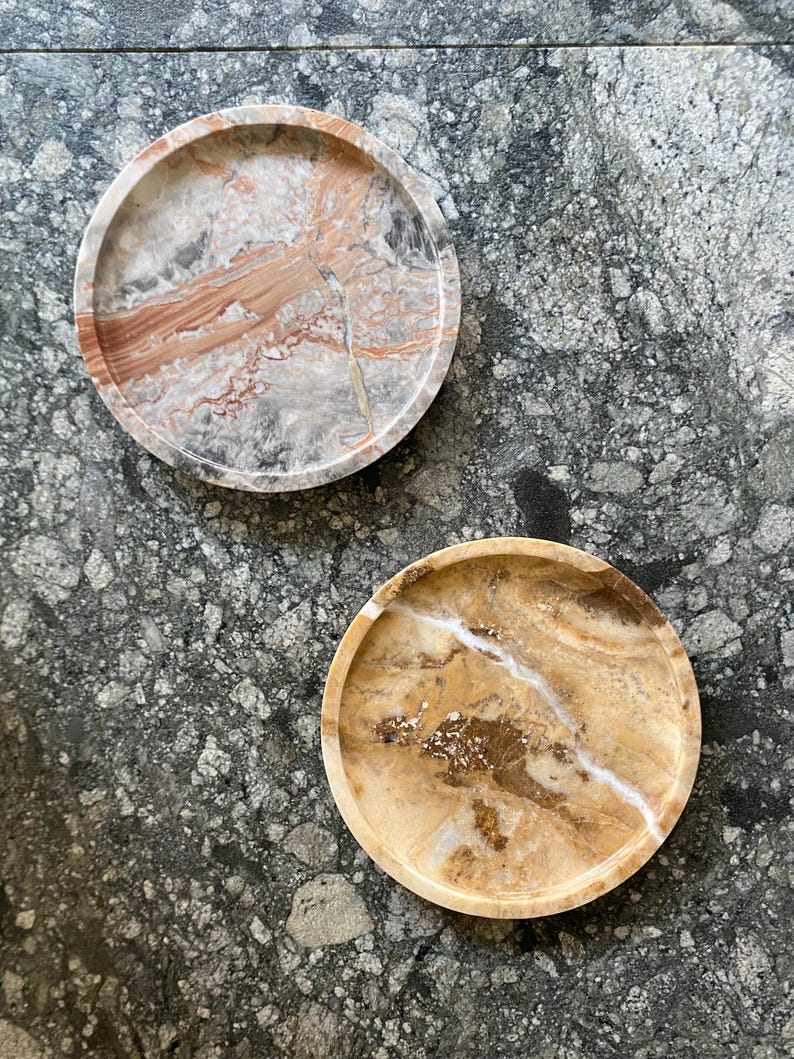 May include: Two circular stone trays with raised edges. The top tray features a marbled pattern of grey, white, and reddish-brown tones. The bottom tray displays a warm amber and golden-brown color palette with white veining and crystalline details.