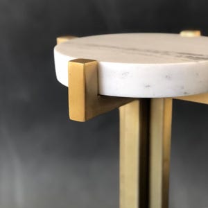 Könnte beinhalten: Nahaufnahme eines kleinen, runden Beistelltisches. Die Tischplatte ist aus weißem Marmor mit grauer Maserung. Die Beine und Stützen sind aus goldfarbenem Metall. Der Tisch hat ein modernes, minimalistisches Design.