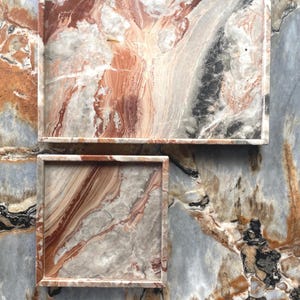 May include: A rectangular tray and a square tray, both crafted from polished stone featuring a marbled pattern of terracotta, cream, gray, and black veins. The trays have raised edges and a smooth, glossy finish, displaying natural stone variations.