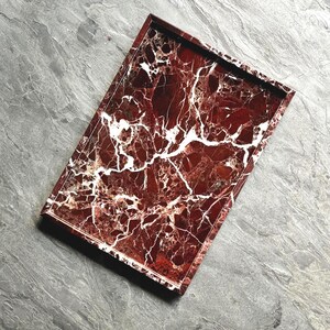 May include: A rectangular tray featuring a deep red and white marble pattern. The surface displays a complex network of white veins running through the dark red stone base, enclosed by a matching red marble frame with raised edges.
