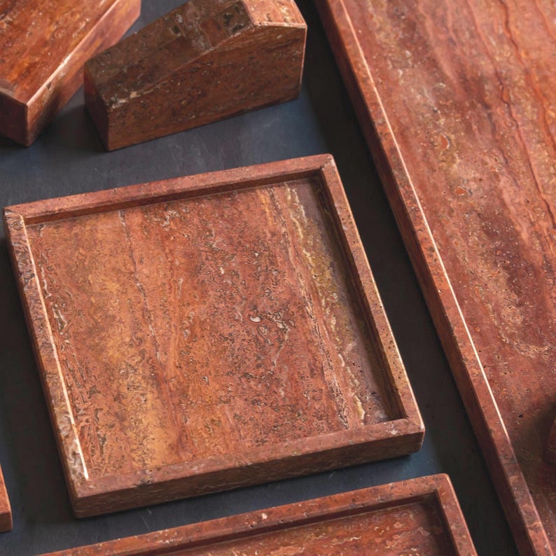 May include: A collection of reddish-brown stone serving trays and blocks. The square tray has a raised edge. The stone has a natural, marbled appearance with variations in color. The items are arranged on a dark surface.