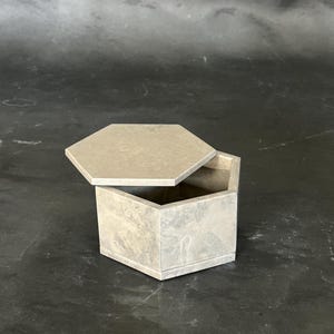 May include: A hexagonal box with a matching lid, both in a light gray color. The lid is slightly ajar, revealing the interior of the box. The box has a geometric design and appears to be made of stone or concrete.