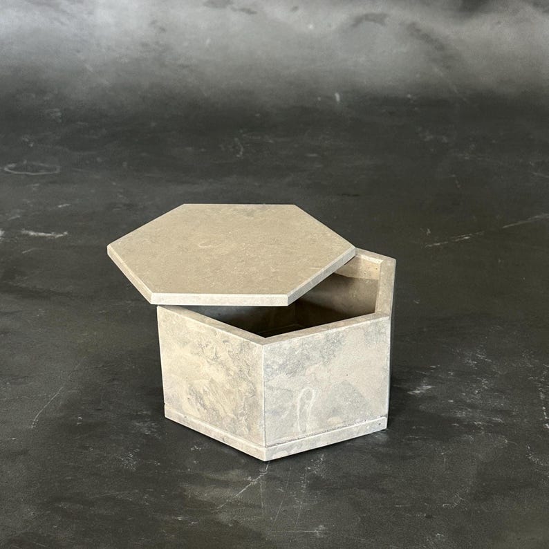 May include: A hexagonal box with a matching lid, both in a light gray color. The lid is slightly ajar, revealing the interior of the box. The box has a geometric design and appears to be made of stone or concrete.