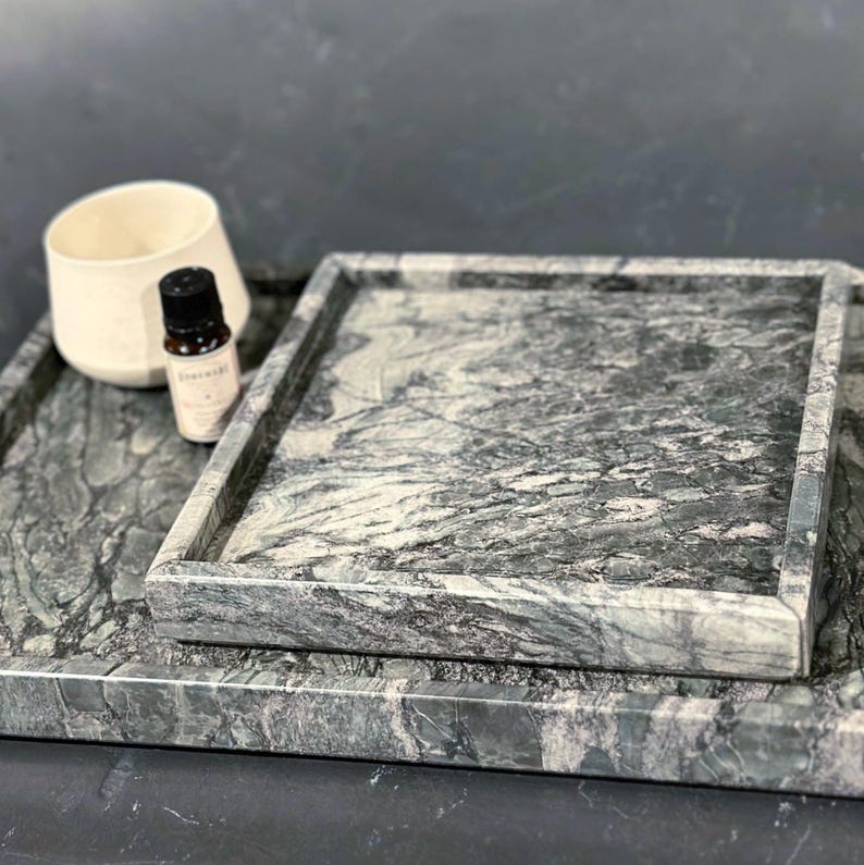 May include: Two marble trays in shades of gray and green, one square and one rectangular, stacked. A small white ceramic cup and a dark bottle with a label are on the trays. The marble has a natural veining pattern.