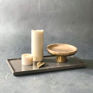K&ouml;nnte beinhalten: Ein rechteckiges Tablett aus dunkelgrauem Stein mit einer hohen zylindrischen cremefarbenen Kerze, einer kleinen runden cremefarbenen Kerze, einem goldenen Blattornament aus Metall und einer beigen Steinschale auf einem Messingfu&szlig;.