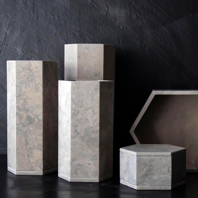 May include: A collection of gray, geometric pedestals and containers with a marbled appearance. The shapes include tall, rectangular columns and hexagonal boxes, all with a stone-like texture. The background is a dark, textured surface.