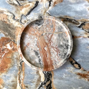 May include: A circular tray crafted from polished stone featuring a complex pattern of swirling gray, white, and deep rust-colored veins. The tray has a smooth, raised rim and a flat base, showcasing the natural variations and textures of the stone.