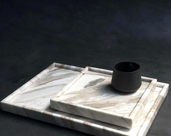 May include: Three white marble trays with brown veining, stacked on top of each other. The top tray has a black ceramic cup on it.