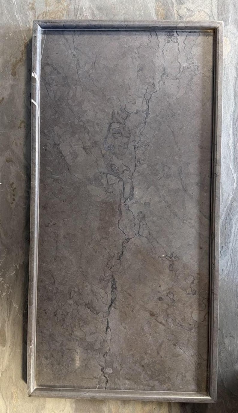 K&ouml;nnte beinhalten: Ein rechteckiges Tablett aus grauem Marmor mit polierter Oberfl&auml;che. Die Fl&auml;che weist durchgehend nat&uuml;rliche, dunkelgraue Aderungen auf, und das Tablett ist von einem erh&ouml;hten, glatten Rand umgeben, der entlang aller vier Seiten verl&auml;uft.