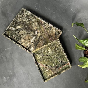 May include: Two rectangular green marble trays with a natural veining pattern. The trays are different sizes and are positioned at an angle to each other. The marble has shades of green, brown, and white. The trays are on a dark gray surface.