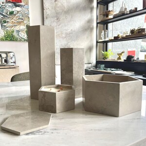 May include: A collection of light gray concrete home decor items, including geometric candle holders, a hexagonal tray, and tall, rectangular pillars. The candle holder has wooden wicks. The items are arranged on a marble surface.