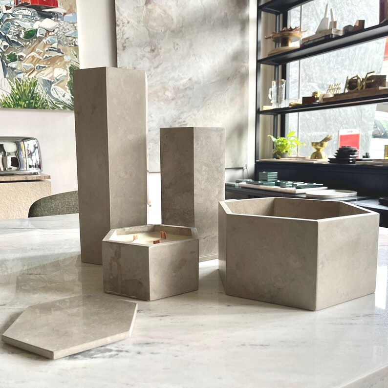 May include: A collection of light gray concrete home decor items, including geometric candle holders, a hexagonal tray, and tall, rectangular pillars. The candle holder has wooden wicks. The items are arranged on a marble surface.