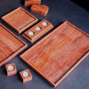 May include: A collection of reddish-brown stone trays and candle holders. The set includes rectangular and square trays, along with candle holders designed to hold small, round candles. The items are arranged on a dark surface.