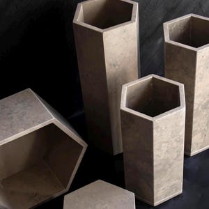 May include: A collection of geometric, hexagonal vases and containers in varying sizes. The vessels are a neutral, mottled gray-brown color, with a matte finish. The arrangement includes both tall, slender vases and shorter, wider containers.