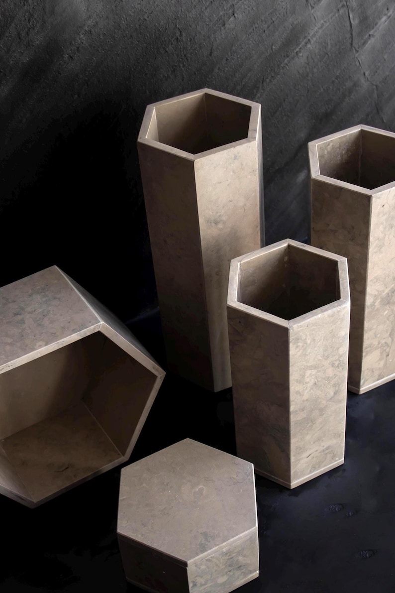 May include: A collection of geometric, hexagonal vases and containers in varying sizes. The vessels are a neutral, mottled gray-brown color, with a matte finish. The arrangement includes both tall, slender vases and shorter, wider containers.