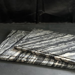 May include: Two rectangular marble cutting boards with a gray, black, and white striped pattern. The boards are stacked, with the top board slightly smaller than the bottom one. The edges have a carved groove.