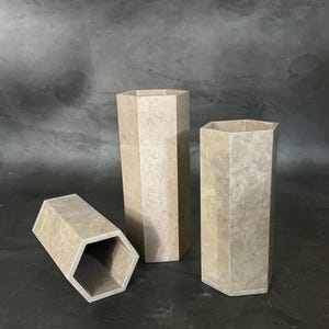 May include: Three beige, hexagonal vases of varying heights. The vases have a marbled appearance and are arranged on a dark surface. One vase is lying on its side, while the other two stand upright.