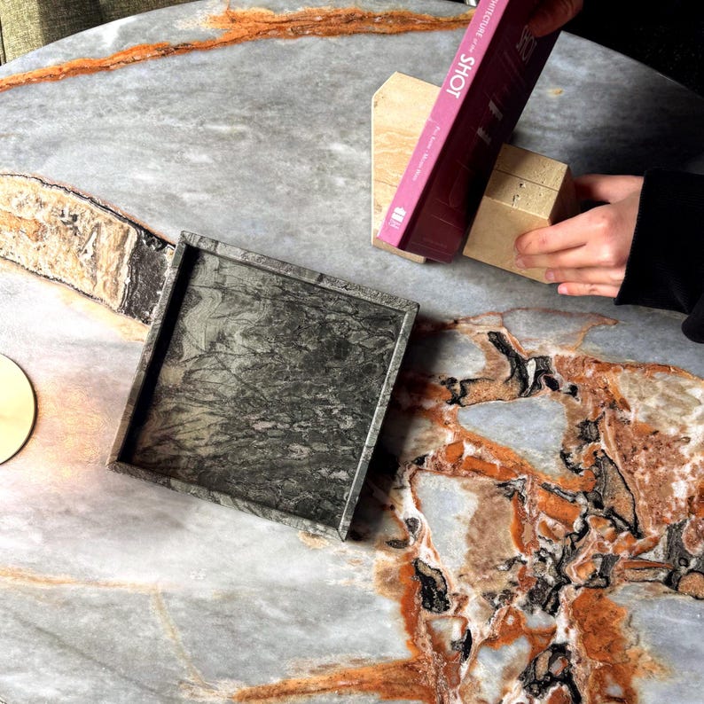 May include: A square, dark green marble tray with a marbled pattern sits on a gray and orange marble table. A book titled "ARCHITECTURE SHOT" and a small, light-colored block are also on the table.