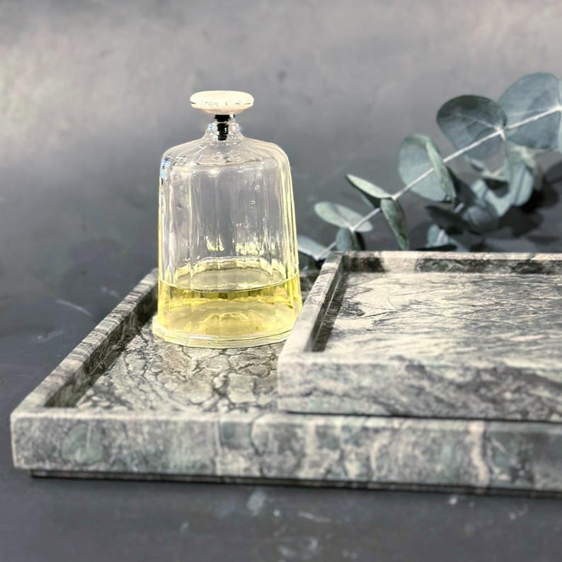 May include: A clear glass cloche with a white-topped stopper, containing a yellow liquid, sits on a gray marble tray. Another marble tray is stacked below. The trays and cloche create a decorative display.