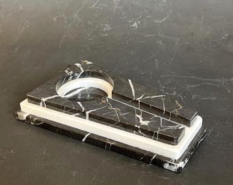 May include: A black marble ashtray with white veining and a white base. The ashtray has a curved opening and a rectangular shape. The design is modern and minimalist, suitable for home decor.