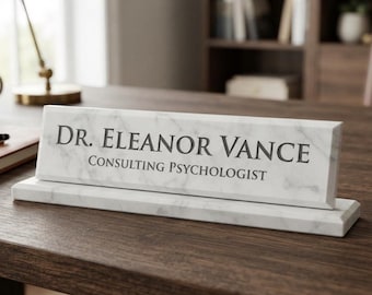 May include: A white marble desk nameplate with grey veining, featuring the engraved text DR. ELEANOR VANCE and CONSULTING PSYCHOLOGIST in a dark grey serif font. The rectangular nameplate rests on a matching marble base on a wooden surface.