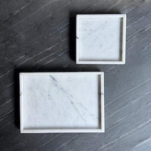 May include: Two white marble trays with grey veining. One tray is square and the other is rectangular. Both trays feature raised edges and a smooth, polished surface. The natural stone displays unique grey patterns across the white base.