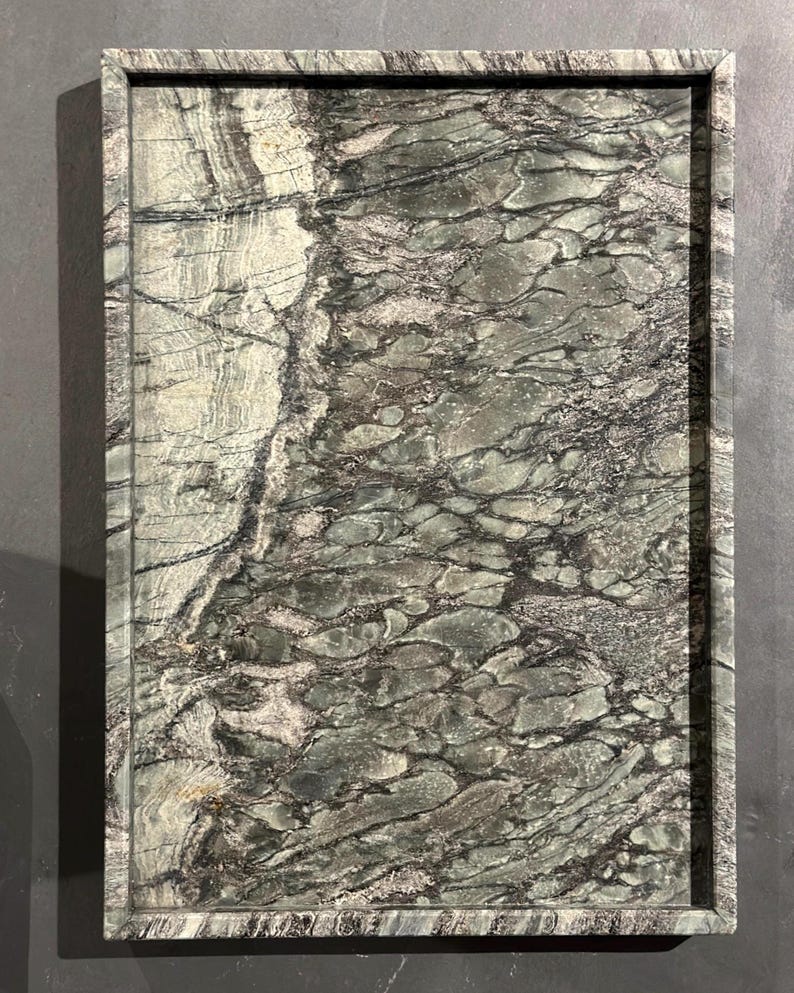 May include: Rectangular serving tray with a natural stone appearance. The tray features a dark gray border and a surface with a swirling pattern of gray, green, and black tones. The tray's design evokes a sense of organic elegance.