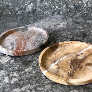 May include: Two circular stone trays with raised edges. One tray features a polished surface with swirling patterns of grey, white, and reddish-brown. The second tray displays a warm, translucent golden-brown finish with prominent white veining.