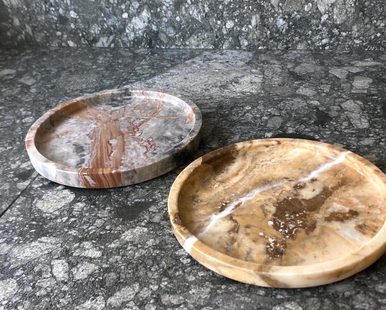 May include: Two circular stone trays with raised edges. One tray features a polished surface with swirling patterns of grey, white, and reddish-brown. The second tray displays a warm, translucent golden-brown finish with prominent white veining.