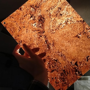 Op de afbeelding: Een vierkant, getextureerd paneel in koperkleur met een gat in de hoek. Het paneel wordt tegen het licht gehouden, waardoor de ingewikkelde oppervlakte details zichtbaar worden. Het algemene effect is een warme, aardse toon.