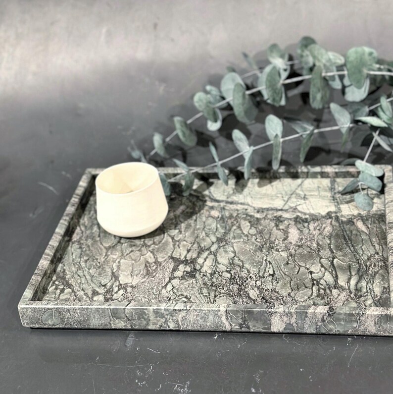 May include: A rectangular marble tray with a green and gray pattern. A small, off-white ceramic cup sits on the tray. Eucalyptus branches are in the background. The tray is approximately 12 inches long and 8 inches wide.