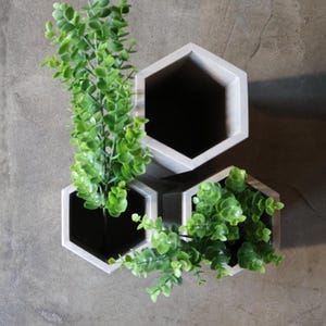 May include: Three hexagonal wall planters in a light gray color, each filled with vibrant green artificial plants. The planters are mounted on a gray concrete wall, creating a modern, minimalist aesthetic.