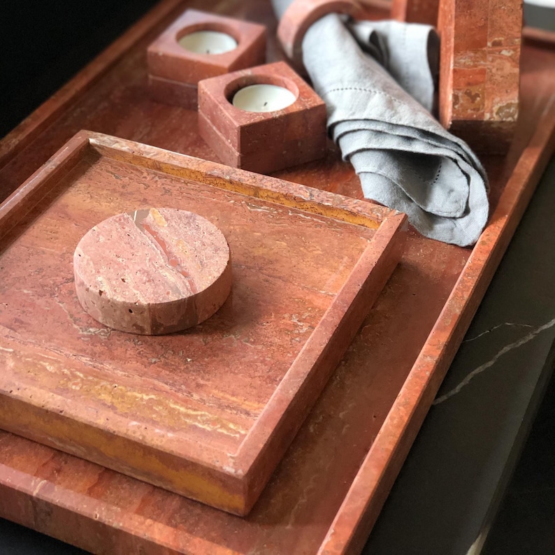 May include: A set of reddish-brown stone trays, candle holders, and a napkin ring. The square trays are stacked, with a round stone piece on top. Two square candle holders with lit candles are visible. A gray linen napkin is folded.