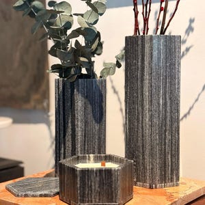 May include: A collection of decorative items, including two tall, gray striped vases holding eucalyptus and red twig branches, and a hexagonal candle with a wooden wick. A matching coaster sits nearby. The items are displayed on a marble surface.