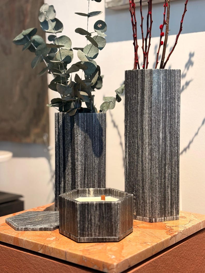 May include: A collection of decorative items, including two tall, gray striped vases holding eucalyptus and red twig branches, and a hexagonal candle with a wooden wick. A matching coaster sits nearby. The items are displayed on a marble surface.