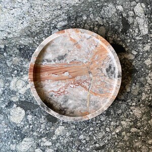 May include: A circular tray featuring a polished stone surface with a natural marbled pattern in shades of terracotta, cream, gray, and white. The tray has a raised, smooth rim and a flat base, showcasing intricate veining throughout the material.