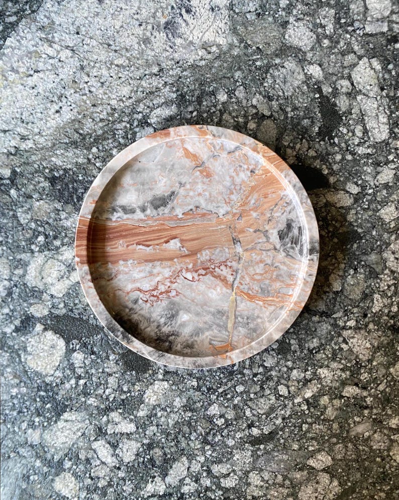 May include: A circular tray featuring a polished stone surface with a natural marbled pattern in shades of terracotta, cream, gray, and white. The tray has a raised, smooth rim and a flat base, showcasing intricate veining throughout the material.