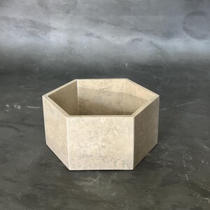 May include: A beige, hexagonal container with a textured surface. The open-top vessel is made of a stone-like material and is suitable for storage or display. The geometric design adds a modern touch.