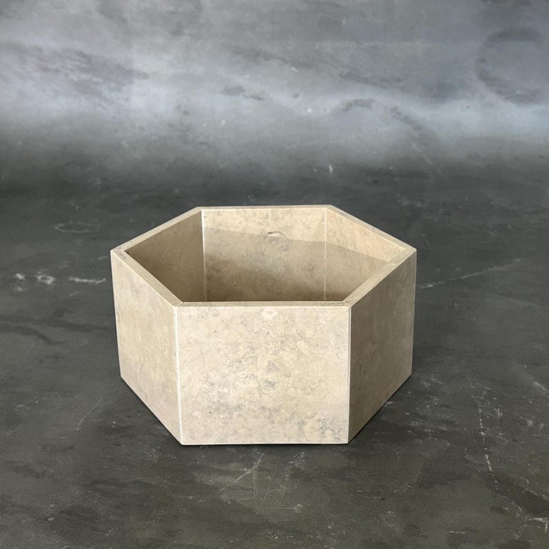 May include: A beige, hexagonal container with a textured surface. The open-top vessel is made of a stone-like material and is suitable for storage or display. The geometric design adds a modern touch.