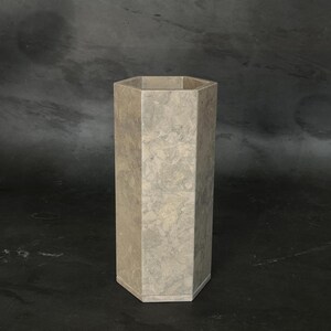 May include: A tall, hexagonal vase with a light gray, marble-like appearance. The vase has a geometric design and a flat base. The neutral color and shape make it suitable for various decor styles. The vase is approximately 24 inches tall.