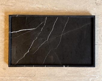 May include: A rectangular black marble tray featuring a smooth surface with distinct white veining patterns throughout. The tray is framed by a thin, raised black border that defines its edges, creating a clean and structured appearance.