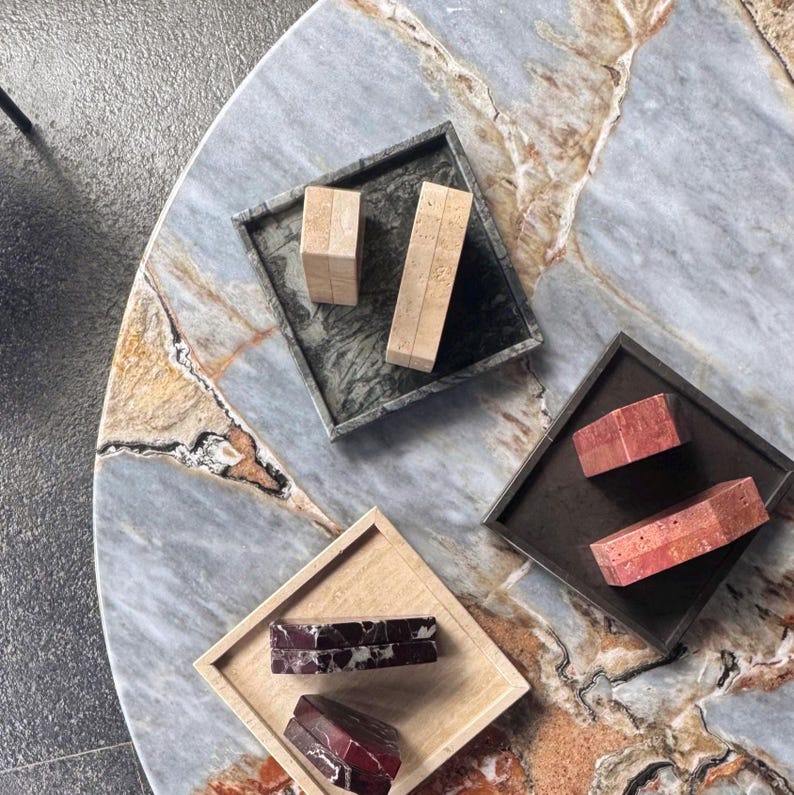 May include: Overhead view of a marble table with three square trays. Each tray holds rectangular stone blocks in varying colors and patterns, including beige, red, and dark green. The table has a blue, gray, and brown marble design.