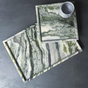 Könnte beinhalten: Zwei rechteckige Serviertabletts und eine kleine runde Tasse aus grün-weißem Marmor. Die Tabletts haben einen erhöhten Rand und ein natürliches, fließendes Muster. Die Tasse hat eine einfarbige, hellgraue Farbe.