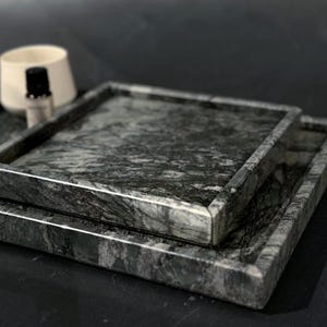 May include: Two square marble trays in shades of gray and green are stacked on top of each other. The trays have a raised edge and a natural stone pattern. A white ceramic container and a small bottle are in the background.