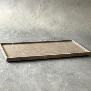 K&ouml;nnte beinhalten: Ein rechteckiges Tablett aus hellbraunem Stein mit einer glatten, polierten Oberfl&auml;che. Das Tablett verf&uuml;gt &uuml;ber einen erh&ouml;hten Rand entlang des gesamten Umfangs und zeigt nat&uuml;rliche Aderungsmuster im Material, die eine subtile, organische Textur erzeugen.
