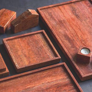 May include: A collection of reddish-brown stone trays and decorative objects. The set includes rectangular and square trays, a candle holder with a lit white candle, and geometric shapes. The stone has a natural, veined pattern.