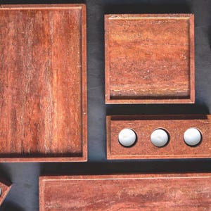 May include: A collection of reddish-brown stone trays and candle holders. The set includes rectangular and square trays, a three-candle holder, and a single candle holder. The items have a natural, textured appearance, and are arranged on a dark surface.