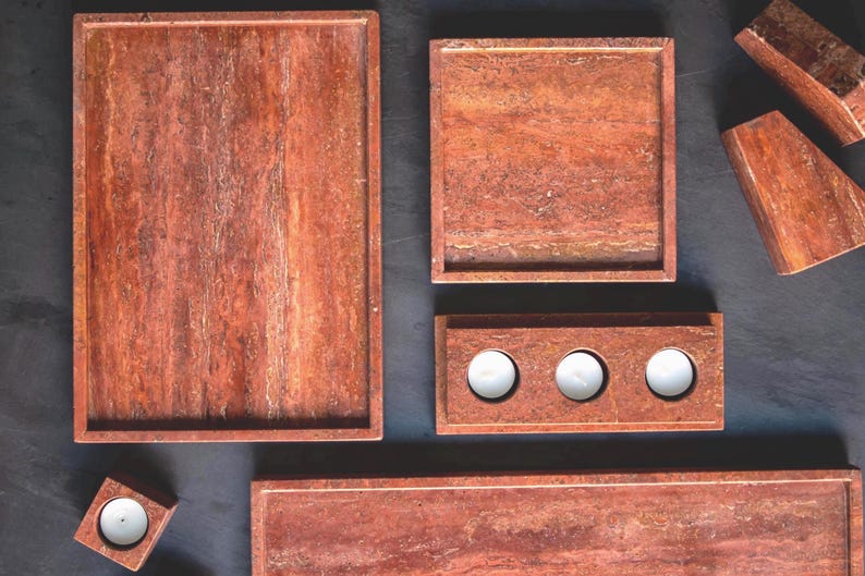 May include: A collection of reddish-brown stone trays and candle holders. The set includes rectangular and square trays, a three-candle holder, and a single candle holder. The items have a natural, textured appearance, and are arranged on a dark surface.